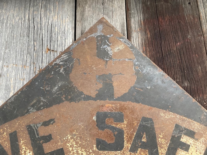 May include: A weathered, triangular metal sign with the word "SAFE" visible. The sign has a black border and a faded, rusty appearance, suggesting age and outdoor exposure. The background is a wooden surface.