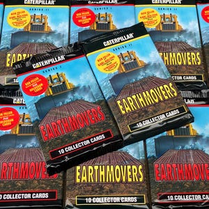 May include: A close-up of a stack of sealed packs of Caterpillar Earthmover trading cards. The packs are black with yellow and red accents. The text on the packs reads "Caterpillar Earthmovers 10 Collector Cards".