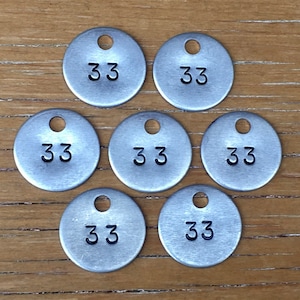 May include: Seven small, round, silver-colored metal tags with the number "33" stamped in black. Each tag has a small hole at the top. The tags are arranged on a wooden surface.