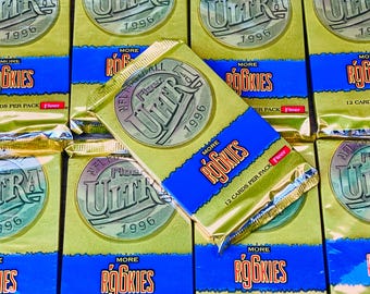 May include: A close-up of multiple sealed packs of 1996 Fleer Ultra NFL trading cards. The packs are gold with a blue and white logo featuring the words "Fleer Ultra" and "1996". The packs also feature the words "More Rookies" and "12 Cards Per Pack".