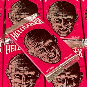 May include: A close-up of a Hellraiser trading card pack. The pack is pink and features a black and white illustration of Pinhead, the main antagonist from the Hellraiser franchise. The text on the pack reads "Hellraiser" and "12 Cards".