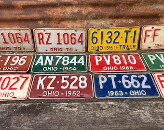 May include: A collection of vintage Ohio license plates with various colors, numbers, and years. The plates include red, white, blue, green, and yellow. The plates are arranged in rows on a wooden surface.