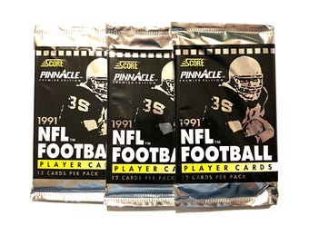 Könnte beinhalten: Drei ungeöffnete Packungen von 1991 Score Pinnacle Premier Edition NFL Football Player Cards. Jede Packung hat einen silbernen Hintergrund mit einem Schwarzweißbild eines Footballspielers, der einen Helm und ein Trikot trägt. Der Text "1991 NFL Football Player Cards" ist in Gelb auf jeder Packung gedruckt.