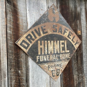 May include: A weathered, diamond-shaped metal sign with the text "DRIVE SAFELY" and "HIMMEL FUNERAL HOME" in black and gold. The sign is mounted on aged wooden planks, showing signs of rust.
