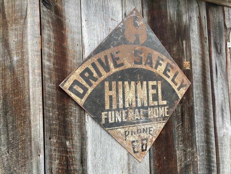 May include: A weathered, diamond-shaped metal sign with the text "DRIVE SAFELY" and "HIMMEL FUNERAL HOME" in black and gold. The sign is mounted on aged wooden planks, showing signs of rust.