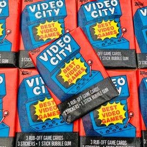 May include: A close-up of several packs of "Video City" trading cards featuring a retro video game console with the text "Best Video Games". Each pack includes 3 rub-off game cards, 3 stickers, and 1 stick of bubble gum.
