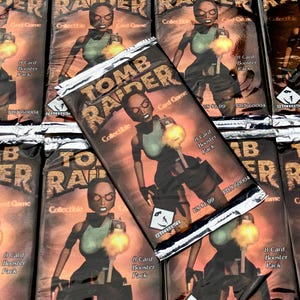 May include: A close-up of multiple sealed packs of Tomb Raider trading cards. The packs are silver with a red, orange, and yellow background. The front of the packs feature a female character holding a gun. The text on the packs reads "Tomb Raider Collectible Card Game".