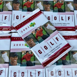 May include: A stack of 2001 Upper Deck Premiere Edition Golf trading card packs. Each pack features a red, white, and black design with the words "GOLF" and "Look for True Rookie Cards of the top players in the game!" The packs are stacked on top of each other, with the top pack showing a picture of Tiger Woods.
