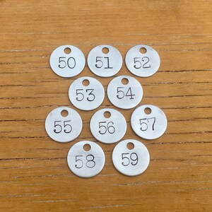 May include: A collection of ten small, silver metal tags, each with a hole at the top and a number from 50 to 59 printed in black. The tags are arranged on a light brown wooden surface.