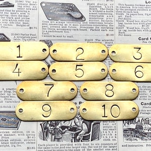 May include: Ten brass number tags, each with a single digit from 1 to 10 in a bold, black font. The tags are oval-shaped with two small holes at each end. The background is a vintage newspaper.