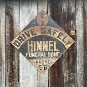 May include: A distressed, diamond-shaped sign with the words "DRIVE SAFELY" at the top. The sign reads "HIMMEL FUNERAL HOME" below. It is mounted on a wooden wall. The phone number "88" is visible. The sign has a weathered appearance.