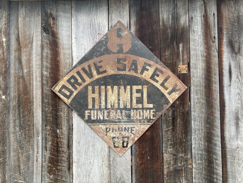 May include: A distressed, diamond-shaped sign with the words "DRIVE SAFELY" at the top. The sign reads "HIMMEL FUNERAL HOME" below. It is mounted on a wooden wall. The phone number "88" is visible. The sign has a weathered appearance.