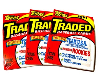 May include: Three Topps 1991 Traded Baseball Cards packs. Each pack is red with yellow and black text, including "Traded Baseball Cards" and "Exclusive". The packs feature "Team U.S.A." and "Promising Rookies".