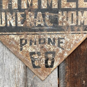 May include: A distressed, triangular metal sign featuring the words "THOMES FUNERAL HOME" in white on black. The sign also displays "PHONE 60" in black. The sign has a rusty, aged appearance, suggesting it is vintage.