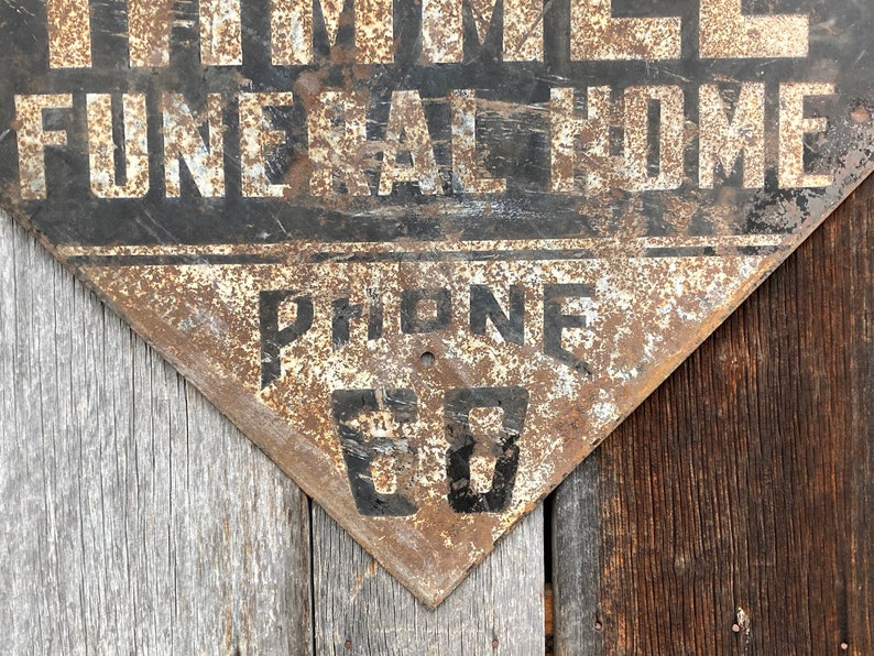 May include: A distressed, triangular metal sign featuring the words "THOMES FUNERAL HOME" in white on black. The sign also displays "PHONE 60" in black. The sign has a rusty, aged appearance, suggesting it is vintage.