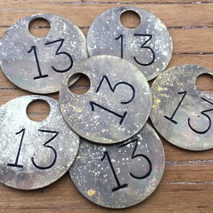 May include: Six brass tags with the number 13 printed on them. The tags are round and have a hole in the center. They are all slightly different shades of brass and have a worn, vintage look.