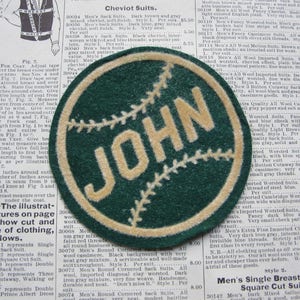 May include: A green and cream felt baseball patch with the name "JOHN" stitched in cream on the front. The patch is circular and has a white stitching detail around the edge.