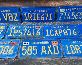 May include: A collection of vintage blue California licence plates with yellow lettering. The plates feature different licence plate numbers, including 4VBZ, 1RIE671, 274565, 1P57416, 1CXP876, 006, 585 AXD, and 1DRZ.