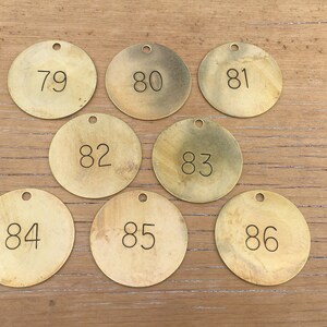 May include: Eight round, brass-colored tags with numbers 79 through 86 printed in black. Each tag has a small hole at the top for hanging. The tags are arranged on a light brown wooden surface.