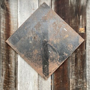 May include: A diamond-shaped metal sign with a rusty, dark surface is mounted on a wooden wall. The sign shows signs of wear, and the wooden planks behind it vary in colour from light grey to dark brown.