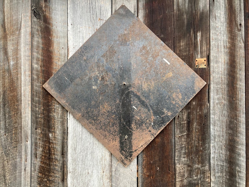 May include: A diamond-shaped metal sign with a rusty, dark surface is mounted on a wooden wall. The sign shows signs of wear, and the wooden planks behind it vary in colour from light grey to dark brown.