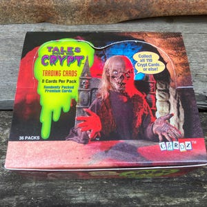 Puede incluir: Una caja de cartas coleccionables de Tales from the Crypt con un personaje espeluznante con ojos rojos y una boca ensangrentada. La caja es roja, blanca y verde con el texto "Tales from the Crypt Trading Cards" y "Collect all 110 Crypt Cards... or else!"
