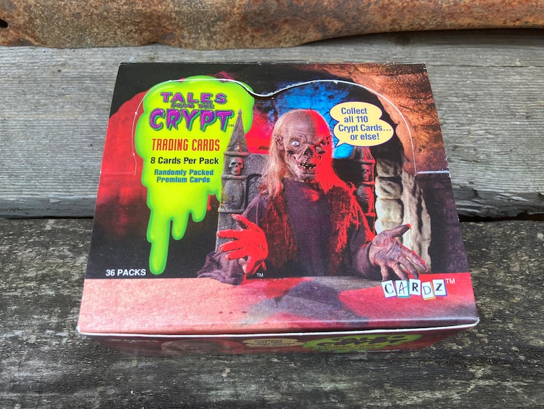 Puede incluir: Una caja de cartas coleccionables de Tales from the Crypt con un personaje espeluznante con ojos rojos y una boca ensangrentada. La caja es roja, blanca y verde con el texto "Tales from the Crypt Trading Cards" y "Collect all 110 Crypt Cards... or else!"