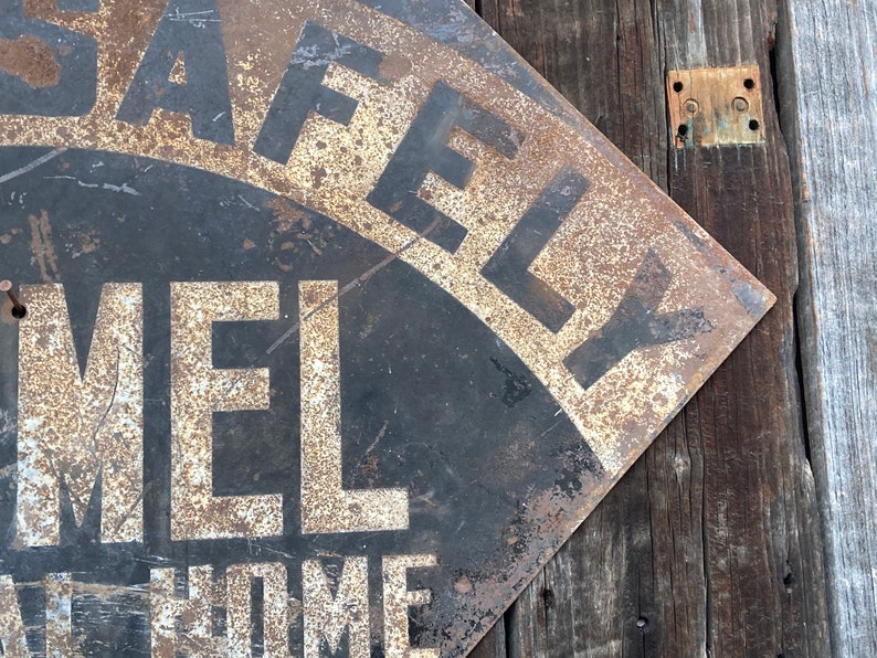 May include: A distressed metal sign with the words "SAFELY" and "MEL" in white lettering on a black background. The sign is mounted on a wooden surface, showing signs of rust and age.