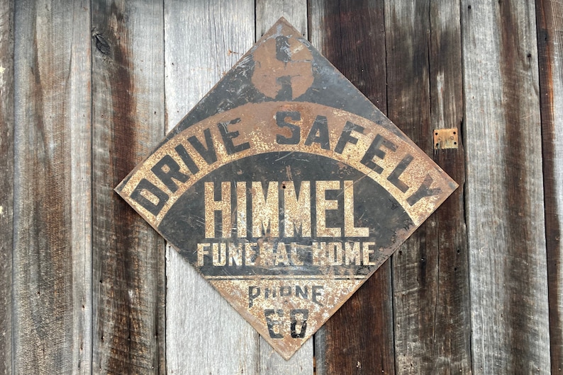 May include: A diamond-shaped, vintage metal sign with the text "DRIVE SAFELY" and "HIMMEL FUNERAL HOME" in black. The sign has a rusty, aged look and is fixed to a weathered wooden wall.