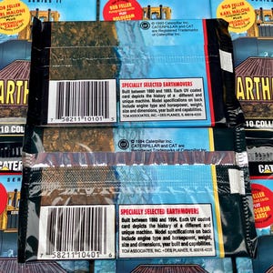 May include: Two unopened packs of Caterpillar Earthmover trading cards. Each pack features a blue and white background with the Caterpillar logo and the text "Earthmovers" in bold white letters. The back of each pack includes a description of the cards and a copyright notice for Caterpillar Inc.