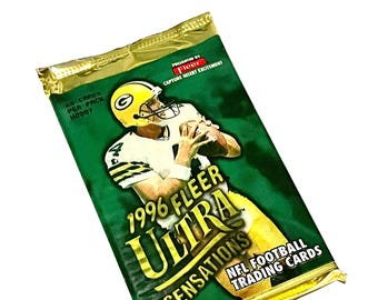 Puede incluir: Un paquete de cartas de fútbol americano 1996 Fleer Ultra Sensations NFL. El paquete es verde y dorado con una imagen de un jugador de fútbol americano con un uniforme verde y dorado. El texto en el paquete dice "1996 Fleer Ultra Sensations NFL Football Trading Cards".