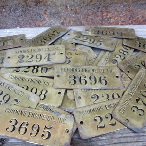 May include: A pile of brass tags with black lettering. The tags are all rectangular and have rounded corners. The text on the tags reads "Cummins Engine Co. Inc." and various numbers.