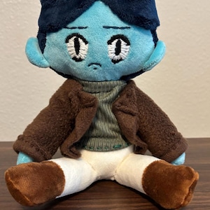 May include: A plush doll with a blue face and dark blue hair, wearing a brown jacket, a green sweater, white pants, and brown boots. The doll has embroidered eyes and a sad expression. The doll is sitting on a wooden surface.