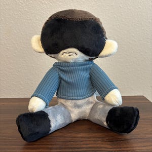 May include: A plush doll with a brown head, a large black eye mask, and a small stitched mouth. The doll wears a ribbed blue long-sleeved shirt and light gray pants with black feet. The doll has cream-colored ears and hands, and it is seated.