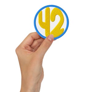 K&ouml;nnte beinhalten: Runder, gestickter Aufn&auml;her mit der gelben Zahl 42 auf wei&szlig;em Grund, umrandet von einem blauen Rand. Der Aufn&auml;her hat eine gen&auml;hte Textur, geeignet f&uuml;r Kleidung oder Accessoires.