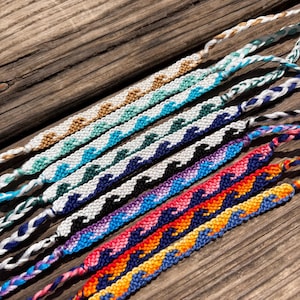 Pode incluir: Uma coleção de seis pulseiras da amizade tecidas à mão com um padrão de ondas repetitivo. Cada pulseira exibe um gradiente de cor único, incluindo fios nos tons de dourado, turquesa, azul escuro, preto, roxo, rosa, laranja e amarelo.