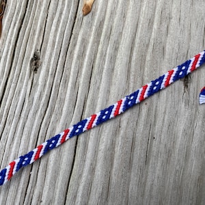 May include: A red, white, and blue friendship bracelet with a diagonal pattern. The bracelet is made with thin string and has a knotted end.