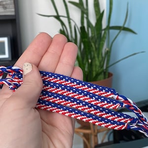 May include: Handmade friendship bracelets in red, white, and blue. The bracelets are made of woven threads, with a diagonal stripe pattern. The bracelets are held in a hand, with a blurred background of a plant and a blue wall.