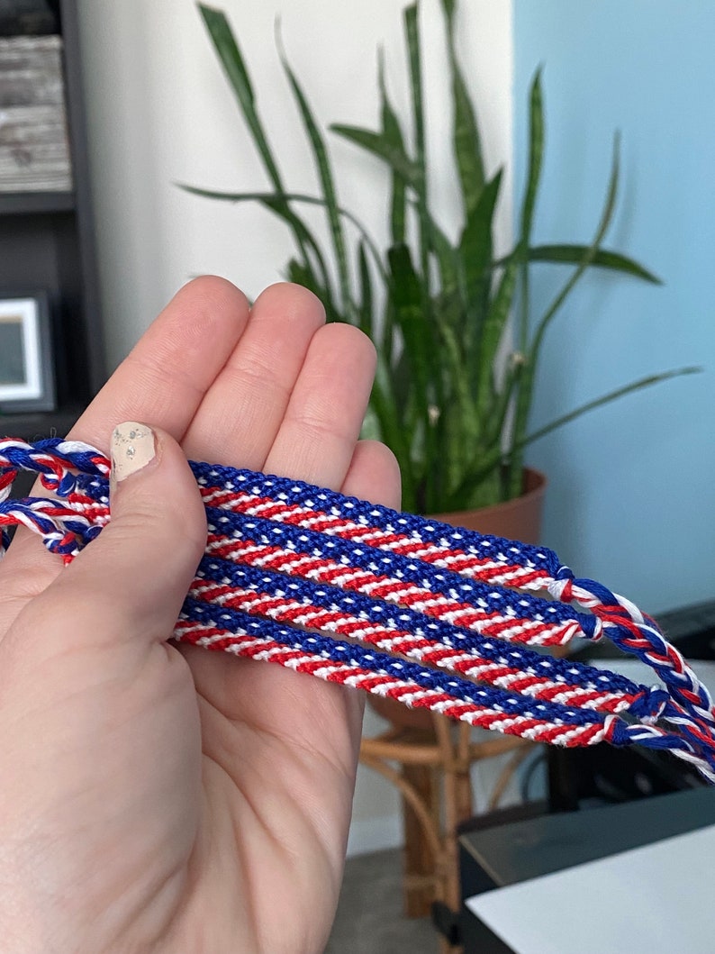 May include: Handmade friendship bracelets in red, white, and blue. The bracelets are made of woven threads, with a diagonal stripe pattern. The bracelets are held in a hand, with a blurred background of a plant and a blue wall.