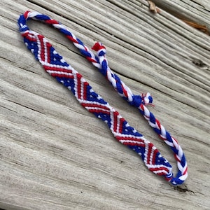 May include: Handmade friendship bracelet featuring a patriotic design. The bracelet is primarily blue, red, and white, with a woven pattern. The bracelet is made of braided threads and is displayed on a wooden surface.