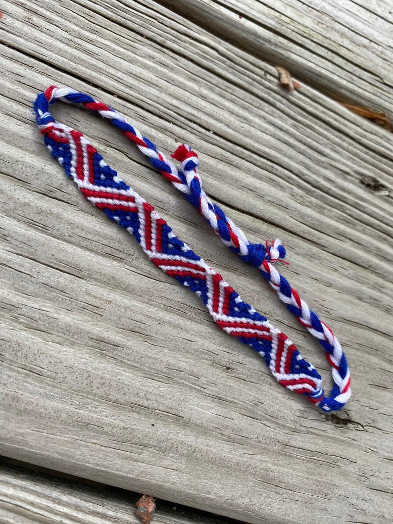 May include: Handmade friendship bracelet featuring a patriotic design. The bracelet is primarily blue, red, and white, with a woven pattern. The bracelet is made of braided threads and is displayed on a wooden surface.