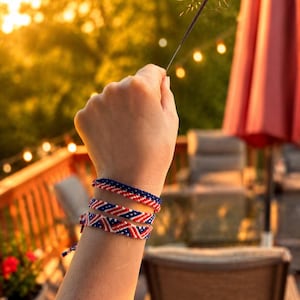 May include: A hand holding a lit sparkler against a blurred outdoor background. The wrist displays three friendship bracelets in red, white, and blue patterns. The bracelets are made of woven threads, with a patriotic theme.