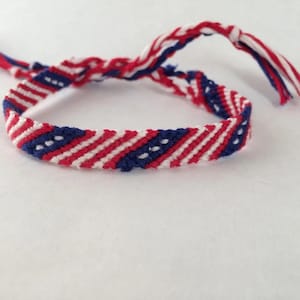 May include: Handmade friendship bracelet in red, white, and blue. The woven bracelet features a striped pattern with small white dots on a blue background. The ends of the bracelet have braided tassels in matching colors.