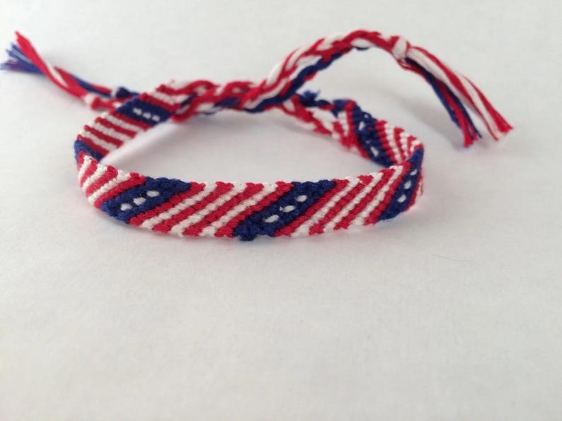 May include: Handmade friendship bracelet in red, white, and blue. The woven bracelet features a striped pattern with small white dots on a blue background. The ends of the bracelet have braided tassels in matching colors.