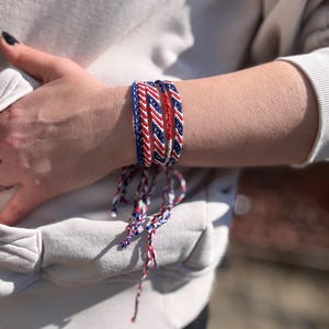 May include: A collection of braided friendship bracelets in red, white, and blue. The bracelets are worn stacked on a wrist. The colors are arranged in a striped pattern. The bracelets are tied with matching colored strings.