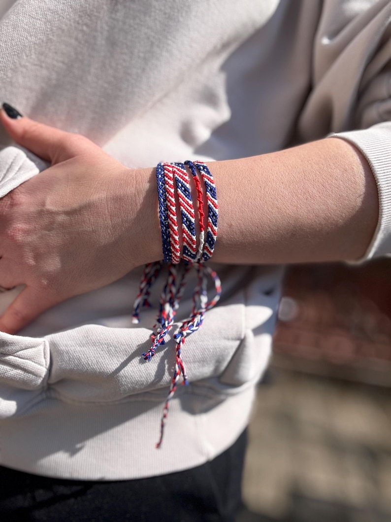 May include: A collection of braided friendship bracelets in red, white, and blue. The bracelets are worn stacked on a wrist. The colors are arranged in a striped pattern. The bracelets are tied with matching colored strings.