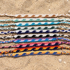 Puede incluir: Una colección de diez pulseras de la amistad tejidas a mano con un patrón de ondas repetitivo. Cada pulsera está elaborada con diferentes combinaciones de colores, incluyendo marrón, turquesa, azul, verde, marino, negro, morado, rosa, rojo y naranja.