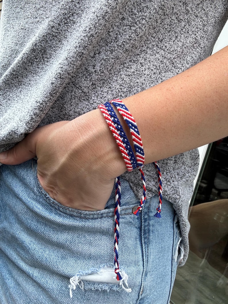 May include: Two braided friendship bracelets in red, white, and blue. The bracelets are worn on a wrist, with the ends of the braids hanging down. The bracelets are a patriotic accessory.