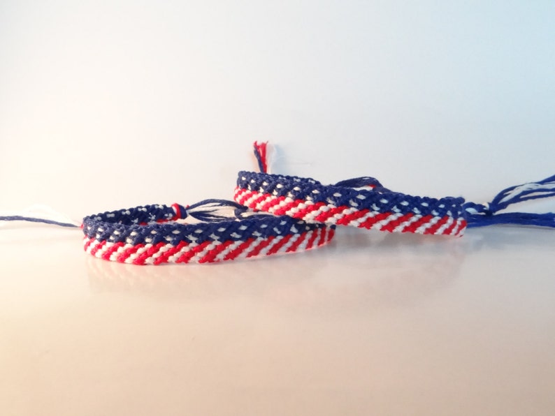 May include: Two handmade friendship bracelets with a red, white, and blue striped pattern. The bracelets are made of woven threads, with a design reminiscent of a flag. The bracelets are displayed on a white surface.