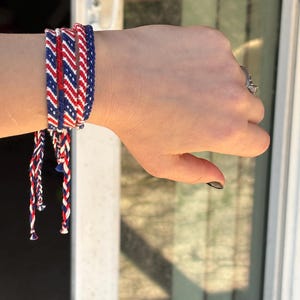 May include: A set of woven friendship bracelets in red, white, and blue. The bracelets are braided with a chevron pattern and tied with long, matching strands. The bracelets are worn on a wrist.